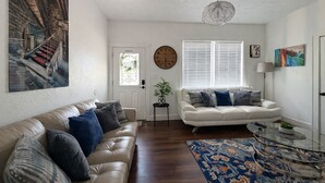 Un salon avec un canapé en cuir beige, un canapé blanc, une table basse en verre, un tapis à motifs, une grande horloge murale et un tableau représentant un escalier.