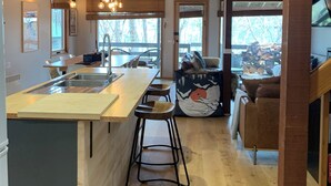 A kitchen with wooden beams, a central island with stools, a dining area with a sofa, and a view of trees through the windows. - Cuff'n'Collar Stable Chalet (Dinner Plain)
