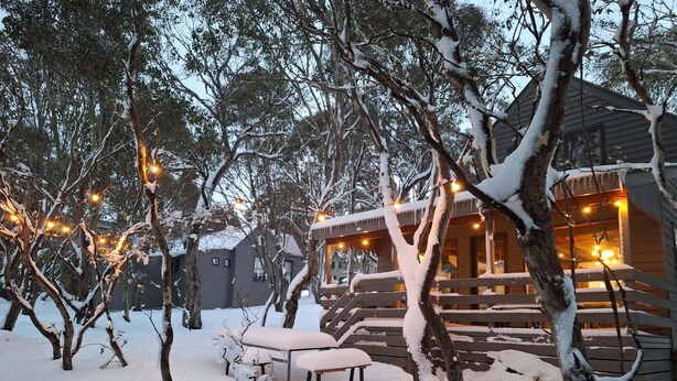 A cabin with a wooden deck and string lights, surrounded by snow-covered trees. - Cuff'n'Collar Stable Chalet (Dinner Plain)