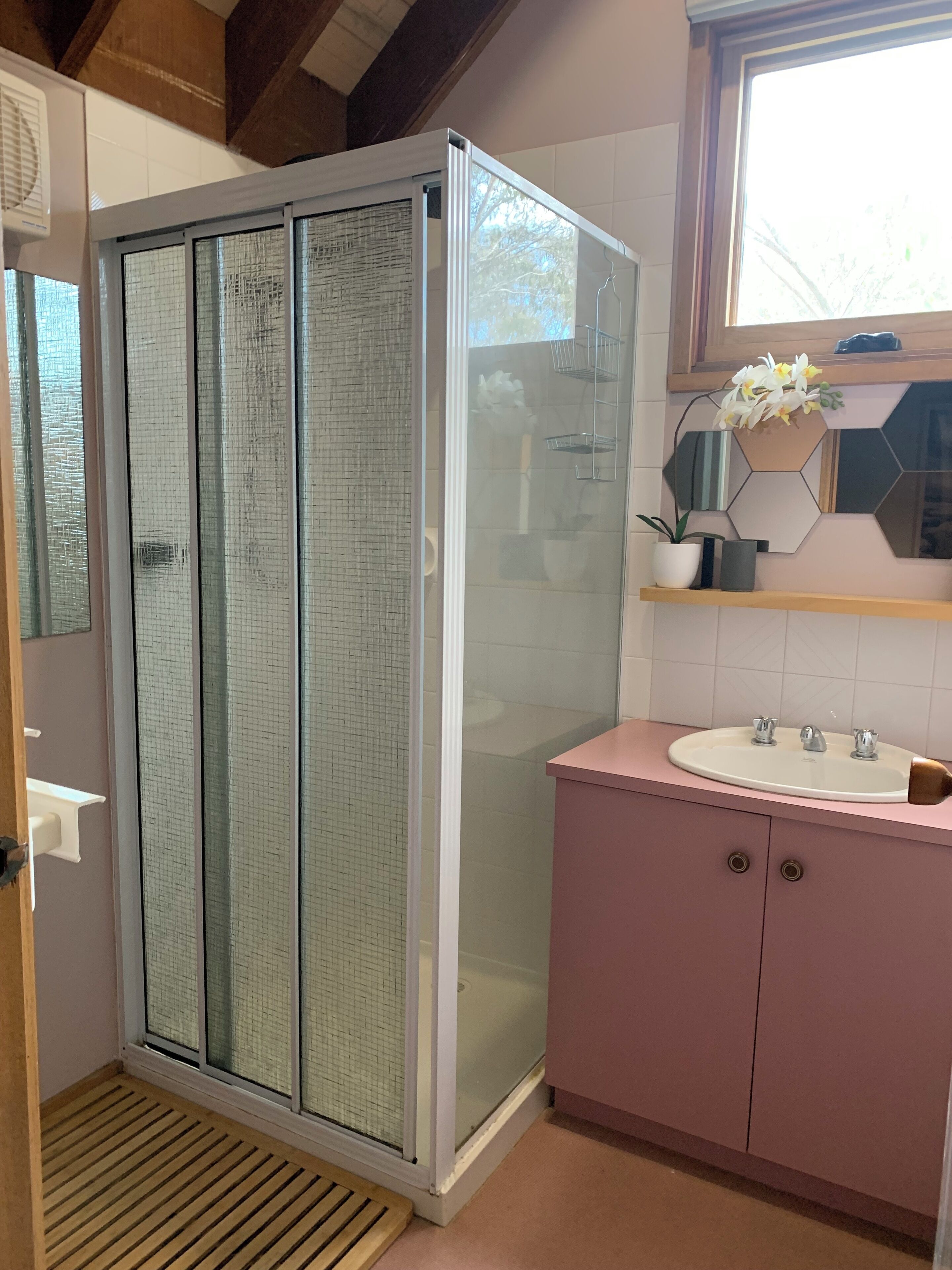 A bathroom with a glass shower enclosure, a pink vanity with a sink, and a decorative shelf with flowers.