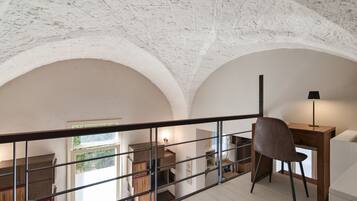 Una stanza con soffitto alto, una scrivania in legno, una sedia e una lampada. Si intravede un balcone con ringhiera e una finestra che lascia entrare luce naturale.
