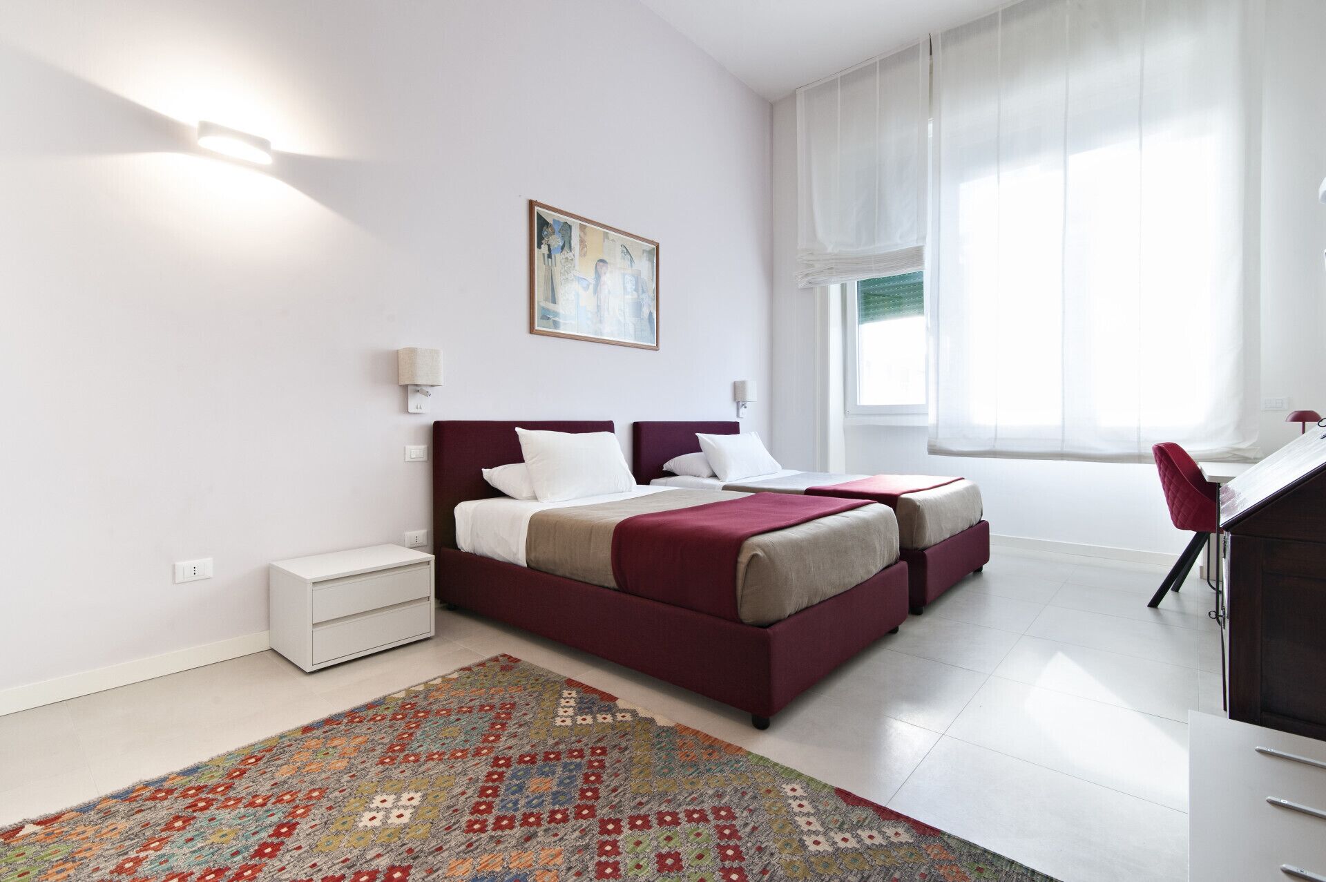 Un dormitorio moderno con una cama grande, mesitas de noche, un escritorio y una ventana con cortinas translúcidas.