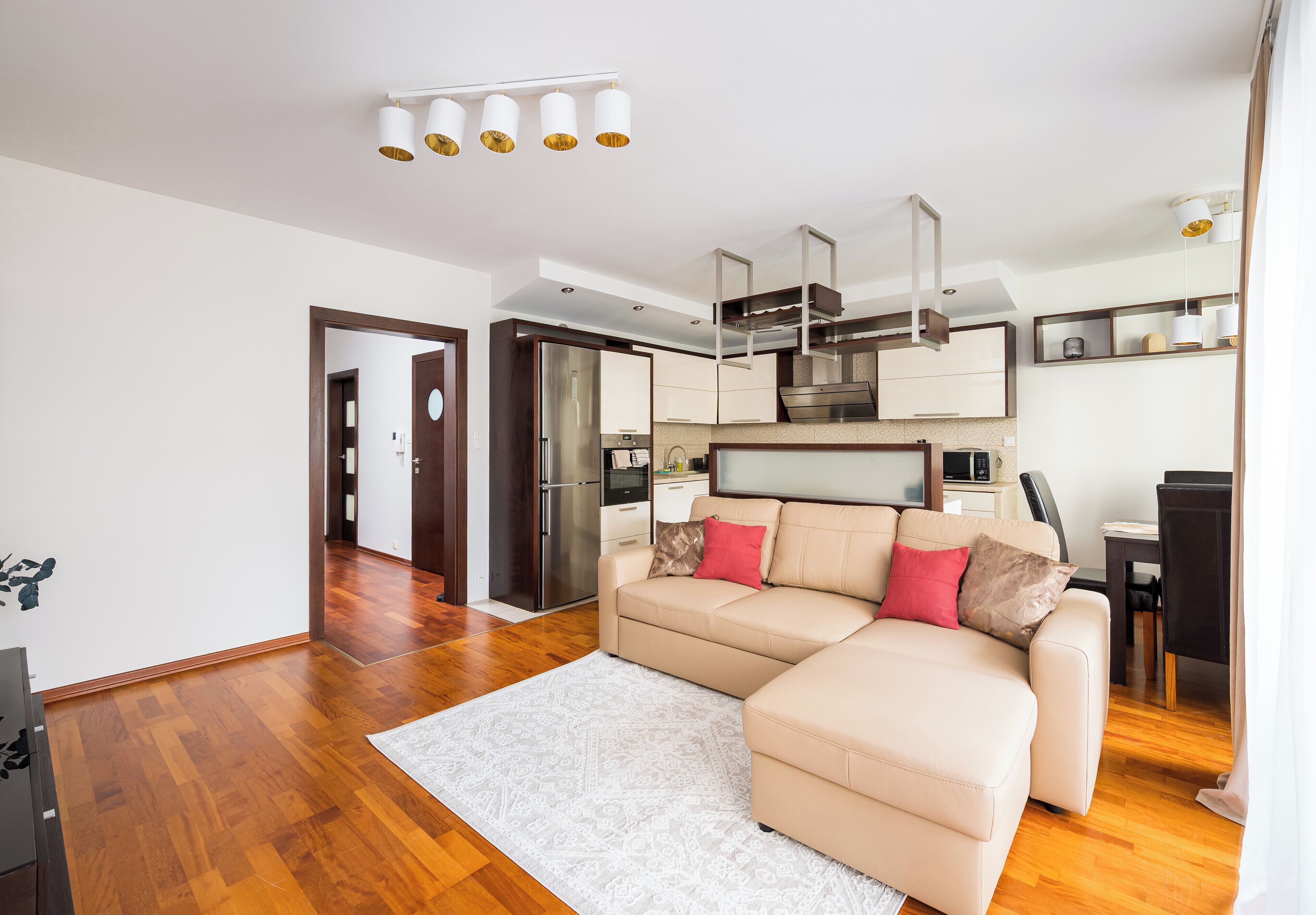 Un soggiorno moderno con un divano angolare beige, un tappeto bianco e un angolo cottura con elettrodomestici in acciaio inossidabile.