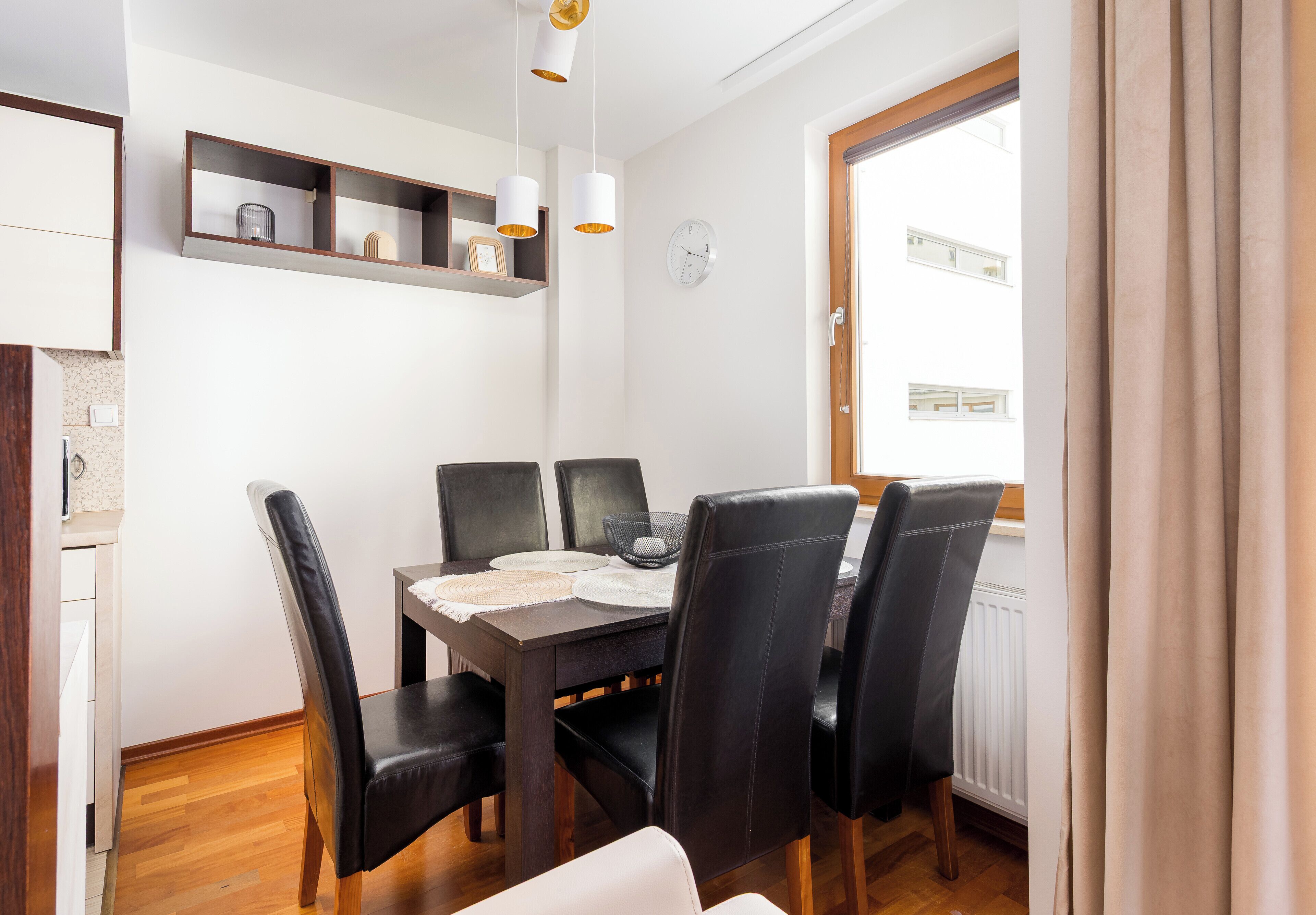 A dining area with a wooden table, black chairs, and pendant lights. There is a clock on the wall and a window with curtains.
