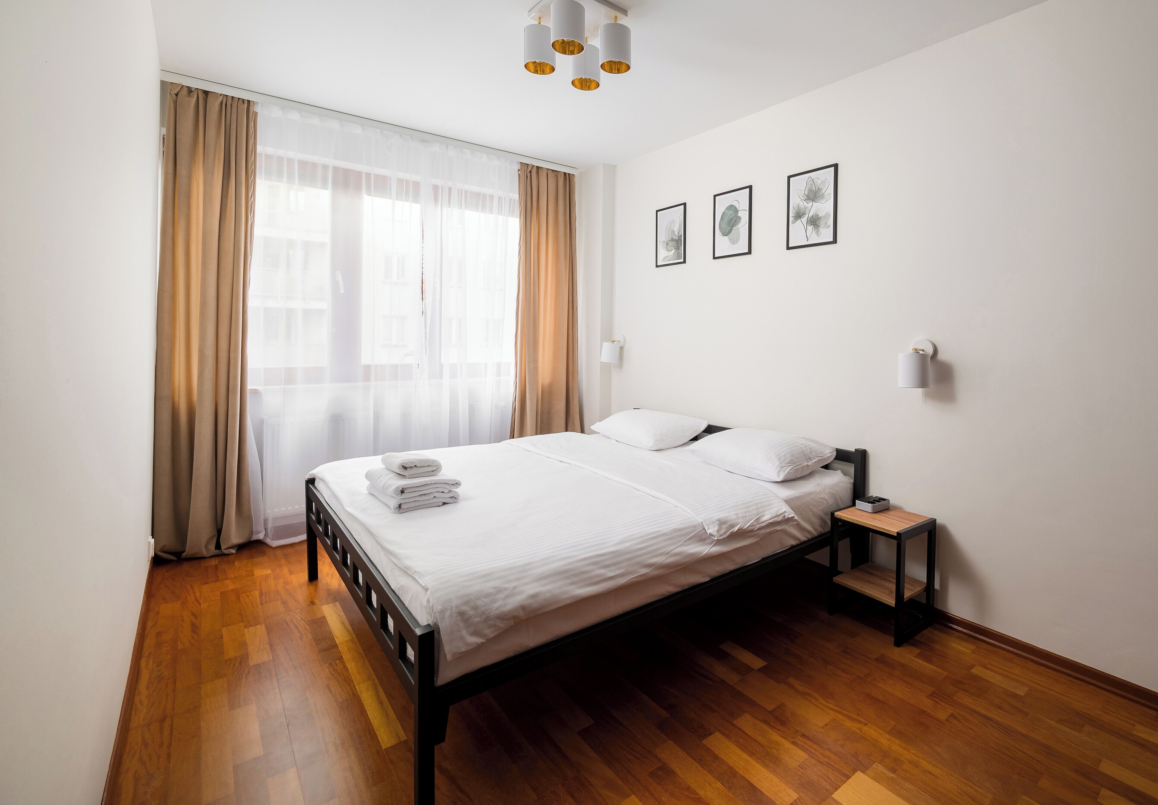 A bedroom with a wooden floor, a bed with white linens, a bedside table, and a window with curtains.