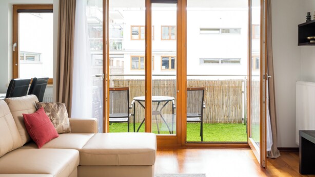 A living room with a sofa, a coffee table, and a sliding glass door leading to a balcony with a view of the neighborhood. - Metro Wilanowska Cosy Apartment (Warszawa)