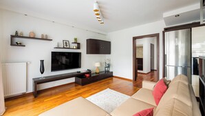 A modern living room with a sofa, a TV, a wooden floor, and a kitchen area visible through an open door. - Metro Wilanowska Cosy Apartment (Warszawa)