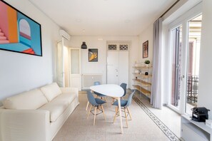 A modern living room with a white sofa, a round table with chairs, a radiator, and a large window with curtains.