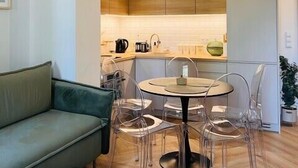 Ein modernes Wohnzimmer mit einem grünen Sofa, einem runden Tisch mit Stühlen, einem Couchtisch, einer Hängeleuchte und einem Küchenbereich im Hintergrund.
