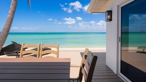 A wooden deck with a table and chairs overlooking a beach with clear turquoise water and a palm tree.