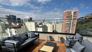 A rooftop terrace with a wooden floor, black leather sofas, and a small wooden table. - Studio with pool in downtown RJ (Santa Teresa Rio de Janeiro)