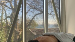 Eine Person liegt in einem Bett, durch das Fenster sind kahle Bäume und ein Gebäude zu sehen.
