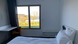 Habitació d'hotel amb un llit, una taula i una finestra que donen vista a vegetació i un edifici.