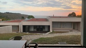 Ein modernes Gebäude mit großen Fenstern und einer flachen Dachkonstruktion, mit Blick auf eine hügelige Landschaft und das Meer.