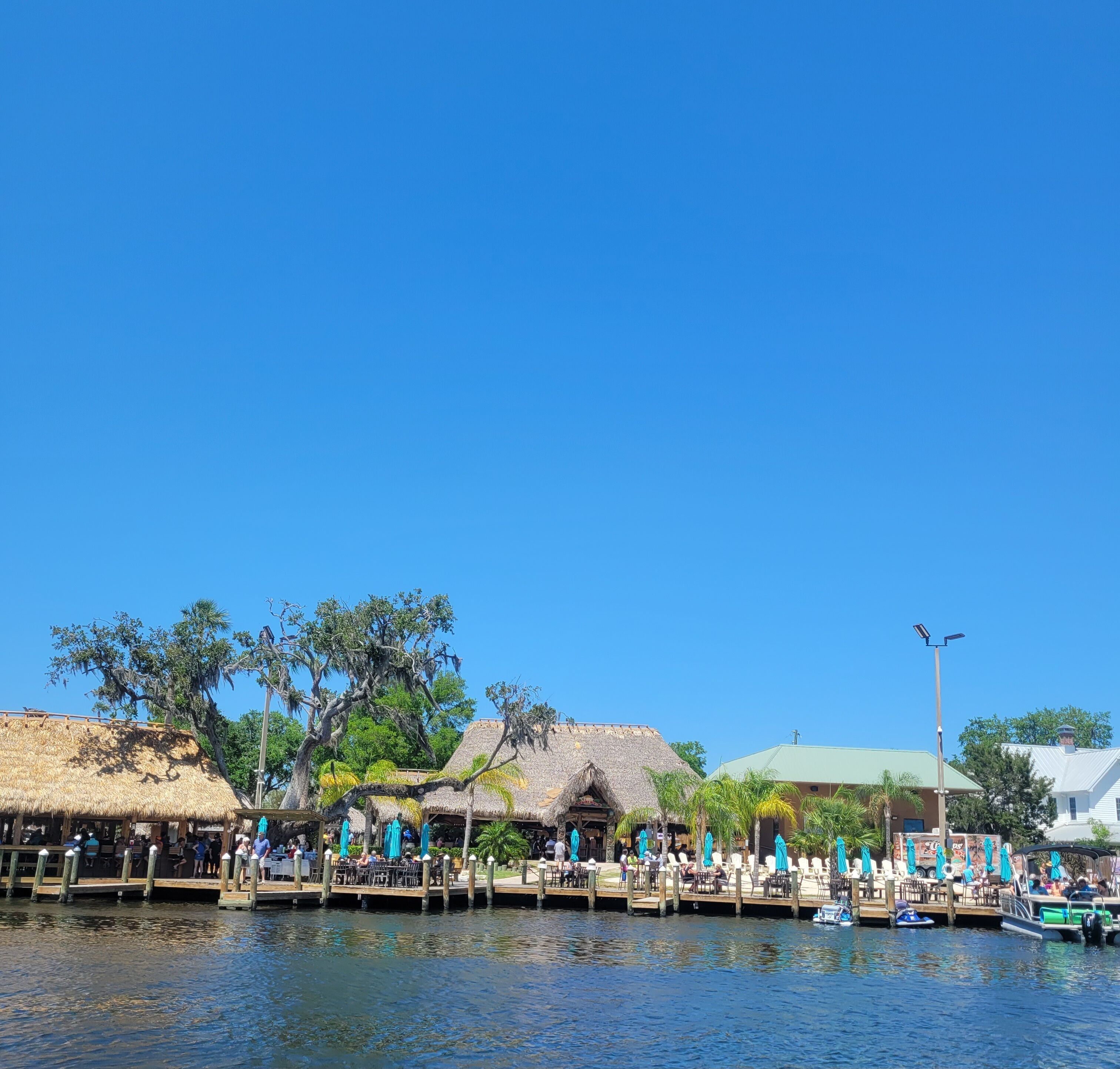 Un restaurante frente al agua con terraza, estructuras de techo de paja y un muelle con barcos.
