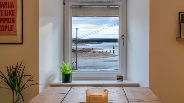 A room with a view of a beach through a window, a candle on a table, and a framed poster on the wall.