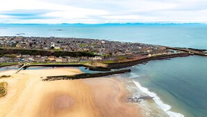 An aerial view of a coastal town with a sandy beach, a rocky breakwater, and a dense cluster of buildings.
