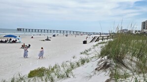 A beach with a pier in the background, sandy terrain, and some vegetation.