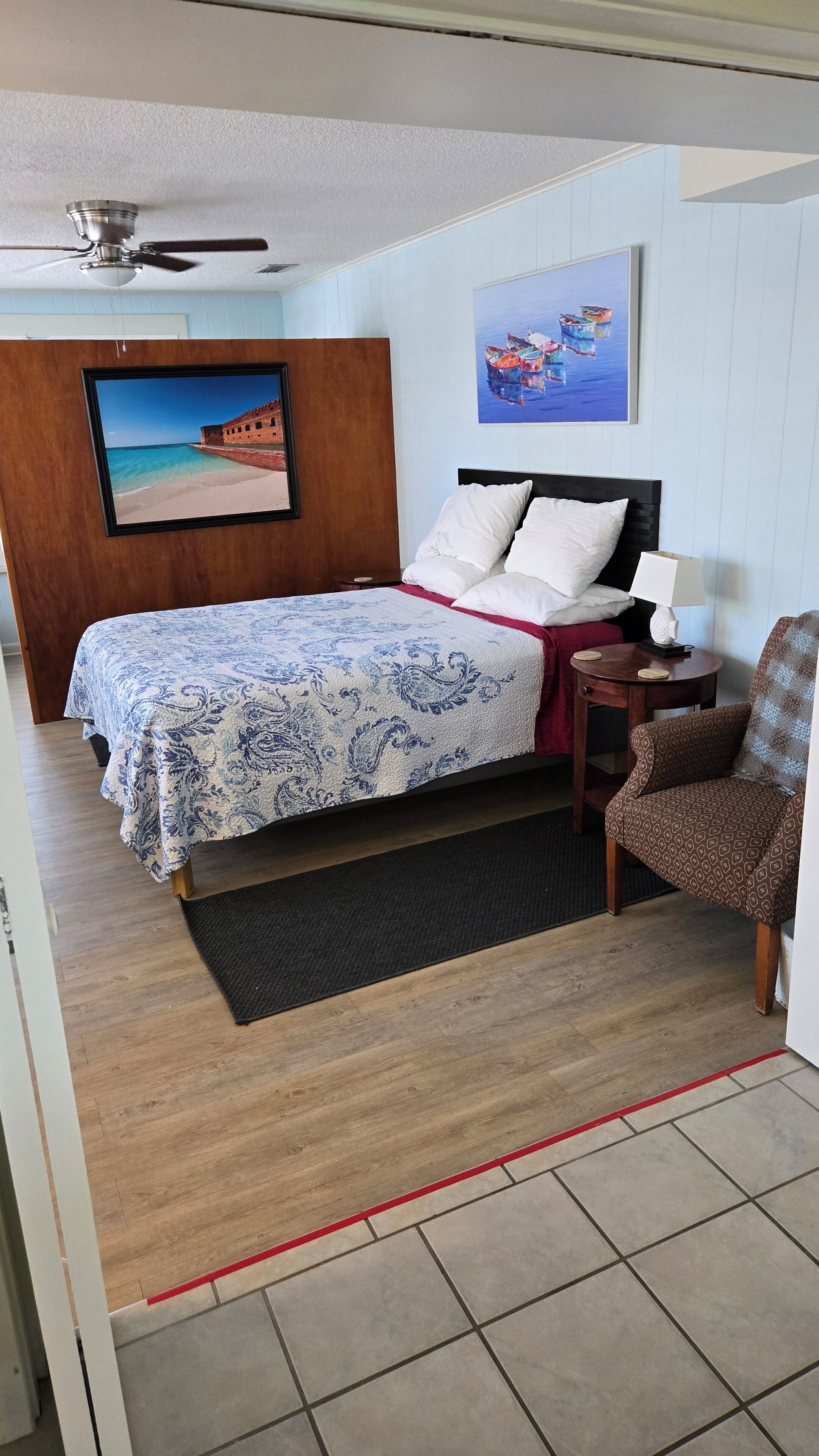 A bedroom with a bed, a nightstand, a chair, a ceiling fan, a painting, and a view of the sea.