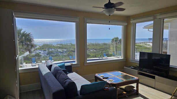 A living room with a sofa, a coffee table, and a TV. There are three large windows offering a view of the beach and ocean.