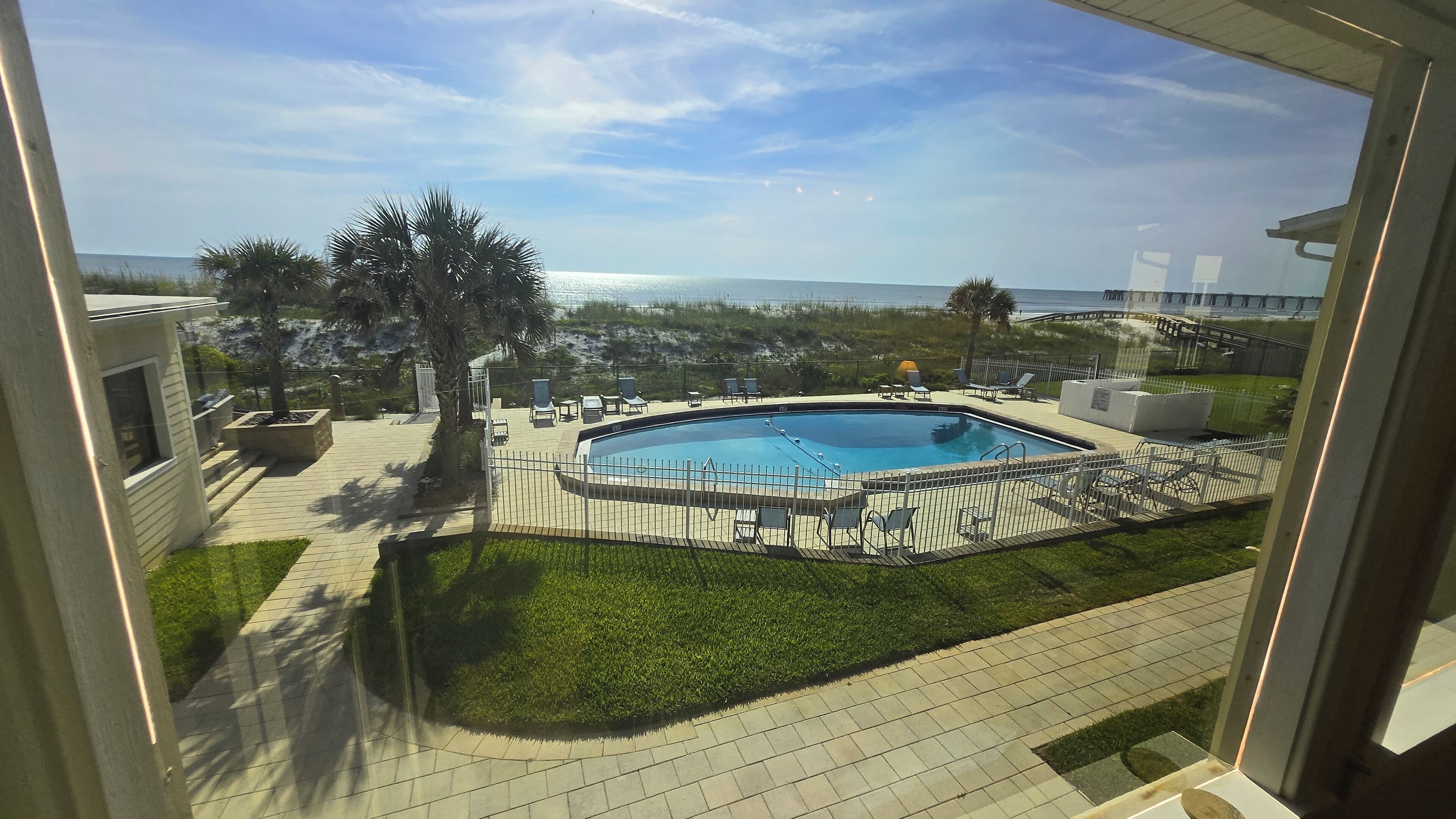 A view of a swimming pool with a surrounding deck, palm trees, and a distant beach.