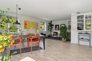 Ein moderner Essbereich mit einem Holztisch und orangefarbenen Stühlen, einem weißen Schrank mit Glastüren und einem großen Fenster mit Blick auf Grünflächen.