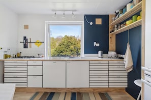 A modern kitchen with white cabinets, a wooden countertop, and a view of trees through a large window.