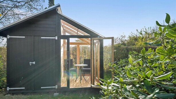 Un cobertizo de madera con puerta corredera de vidrio, un pequeño patio con mesa y sillas, y un jardín con follaje verde.