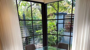 Een eetgedeelte met een glazen tafel, omringd door vier stoelen, grote ramen met gordijnen en uitzicht op groen.