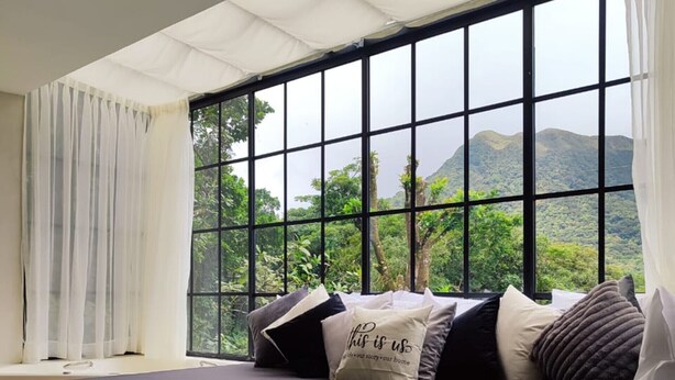 Una habitación con un ventanal grande, una cama con muchas almohadas y vistas a la vegetación y montañas.