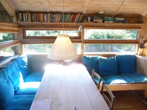 A wooden cabin interior with a dining table, blue upholstered seating, bookshelves, and large windows.