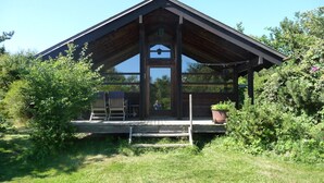 A cabin with a porch, wooden exterior, and a large window. - Cozy cottage close to the beach (Liseleje)