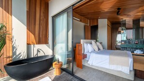 A modern bedroom with a large bed, a glass-enclosed bathroom with a black bathtub, and a wooden floor.