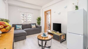 Una sala de estar moderna con un sofá gris, una mesa de centro de madera, un televisor de pantalla plana y un refrigerador blanco.