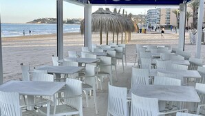 A beachside café with white plastic chairs and tables, a thatched umbrella, and a view of the beach and ocean. - APARTBEACH PARADIS PARK VUE SUR LA PLAGE (La Pineda)