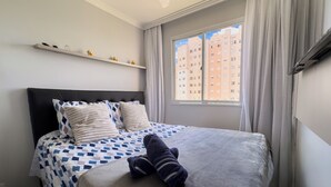 Ein ordentlich bezogenes Bett mit einer gemusterten Tagesdecke, ein weicher Teddybär und ein Fenster mit Vorhängen.