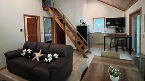 Een ruime woonkamer met een donkerbruine bank, een houten trap en een keuken met een eettafel en barstoelen.