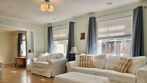 A spacious living room with a beige sofa set, a matching ottoman, and a wooden floor. - House Acadia at Gallagher`s Travels (Bar Harbor)