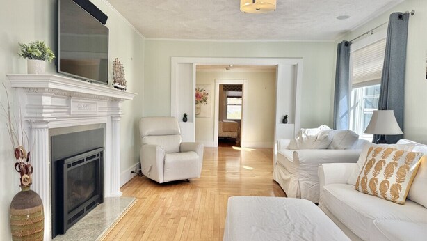 A spacious living room with a fireplace, white furniture, and a television mounted above it. - House Acadia at Gallagher`s Travels (Bar Harbor)
