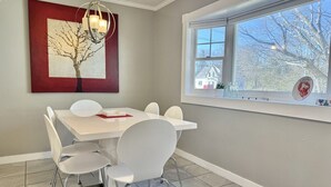 A dining area with a white table and chairs, a chandelier, a framed artwork of a tree, and a window with a view of trees and a house. - House Acadia at Gallagher`s Travels (Bar Harbor)