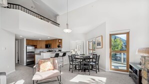 A spacious living room with a high ceiling, a dining area with a round table, and a kitchen with modern appliances. - 2BR + Loft + Sofabed | Tyndall Stone Lodge | Epic Village Location | Mountain Views | Pool & Hot Tub (Whistler)