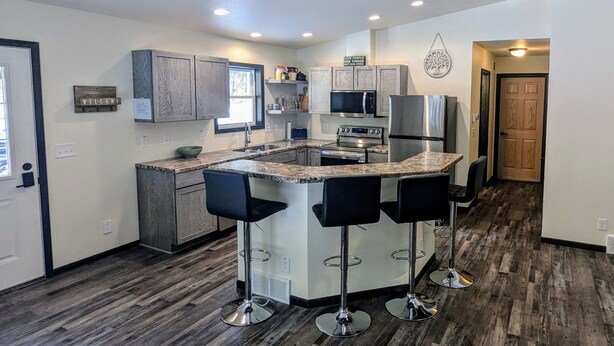 A modern kitchen with a central island, bar stools, stainless steel appliances, and a ceiling fan. - Charming quiet 2-bedroom house set in a cedar forest in welcoming Fish Creek (Fish Creek)