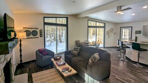 A spacious living room with a large gray sofa, a small armchair, and a coffee table. There are large windows providing a view of a snowy outdoor area. - Charming quiet 2-bedroom house set in a cedar forest in welcoming Fish Creek (Fish Creek)