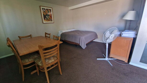 A small room with a wooden table and chairs, a bed with a gray blanket, a standing fan, and a lamp on a cabinet. - Family Suite - Economy (Garden City)