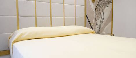 A bed with a white mattress and a yellow pillow in a room with a white padded headboard and a wall-mounted lamp.