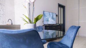 Un espace repas moderne avec une table en verre, des chaises bleues, un téléviseur posé sur un meuble, une plante en pot et une décoration minimaliste.