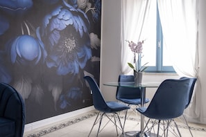 Uma área de refeições com mesa de vidro, cadeiras azuis e um mural floral na parede.