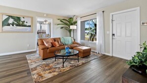A living room with a brown leather sofa, a glass coffee table, and a patterned rug. There is a large window with white curtains, a door, and a plant on the right side. - 4BR, 2BA with Heated Pool, BBQ, Fire pit in Port St. Lucie (Port St. Lucie)