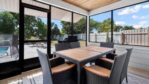 An outdoor patio with a wooden table and wicker chairs, large windows, and a view of a backyard with a barbecue grill. - 4BR, 2BA with Heated Pool, BBQ, Fire pit in Port St. Lucie (Port St. Lucie)