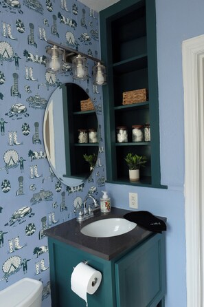 Ein Badezimmer mit blauen Wandmustern, einem weißen Waschbecken, einem Spiegel und einem Regal mit Gläsern und einer Pflanze.